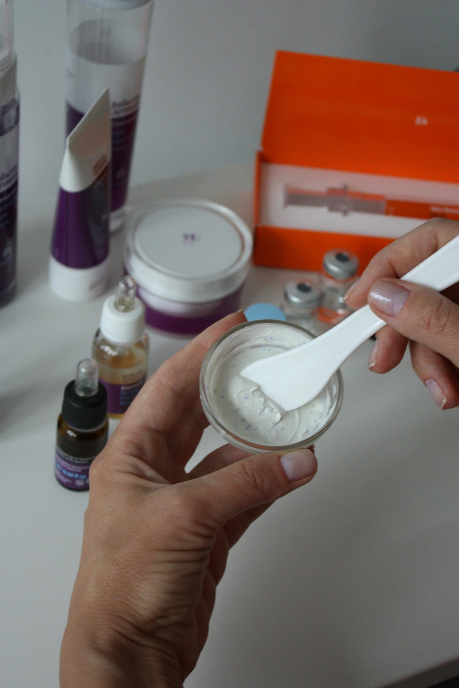 zipwp image 9316658 Close-up of hands mixing beauty product in glass jar, surrounded by cosmetics on white background.